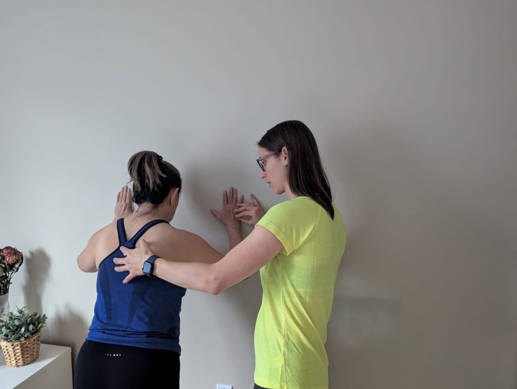 A physiotherapist is standing next to a patient completing a wall push up. The physiotherapist’s hands are on her spine and hand to ensure the patient is completing the exercise appropriately.