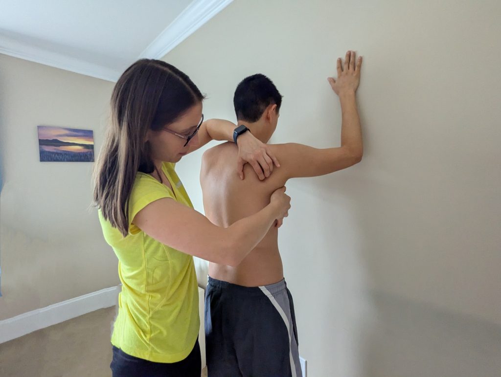 A physiotherapist manually adjusts a patient’s ribcage and shoulder while the patient leans against a wall with one arm raised, focusing on posture and alignment.